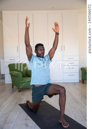 African american man stretching into lunge on black yoga mat in living room in athletic wear African american man stretching into lunge on black yoga mat in living room in athletic wear 140566072