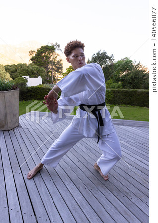 African american woman practicing karate on deck near planter wearing white gi and black belt African american woman practicing karate on deck near planter wearing white gi and black belt 140566077