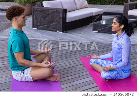 Diverse friends meditating on pink and purple mats on wooden deck patio near sofas Diverse friends meditating on pink and purple mats on wooden deck patio near sofas 140566078