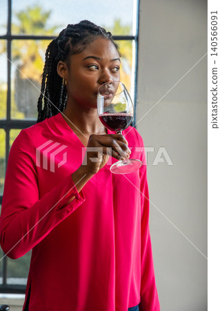 African american woman wearing pink blouse holding wine glass by grid window African american woman wearing pink blouse holding wine glass by grid window 140566091