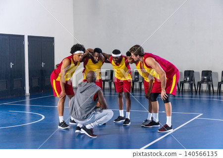 Diverse male basketball team huddling around coach on court, wearing yellow and red jerseys Diverse male basketball team huddling around coach on court, wearing yellow and red jerseys 140566135