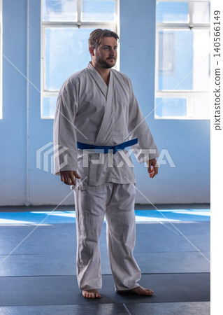Male karate student standing barefoot in white gi blue belt in dojo on mats near windows Male karate student standing barefoot in white gi blue belt in dojo on mats near windows 140566149