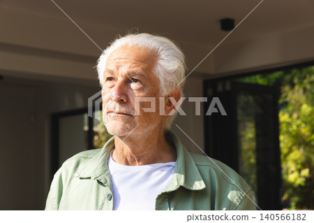 Senior man wearing casual shirt standing in living room facing sliding doors revealing lush garden Senior man wearing casual shirt standing in living room facing sliding doors revealing lush garden 140566182