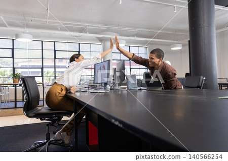 Diverse coworkers in business attire high fiving over desk at modern office with monitor, laptop Diverse coworkers in business attire high fiving over desk at modern office with monitor, laptop 140566254