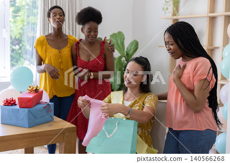 Diverse female friends gathering in home living room and opening pink baby onesie from gift bag Diverse female friends gathering in home living room and opening pink baby onesie from gift bag 140566268