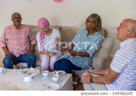 Diverse friends laughing while leaning on sofa in living room around table with cups, cake Diverse friends laughing while leaning on sofa in living room around table with cups, cake 140566285
