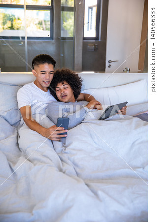 Diverse male couple reclining on bed in bedroom with gray duvet holding smartphone and tablet Diverse male couple reclining on bed in bedroom with gray duvet holding smartphone and tablet 140566333