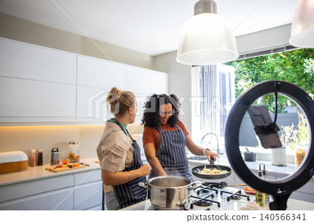 Diverse female friends wearing aprons lifting frying pan, holding pot on gas stove in kitchen Diverse female friends wearing aprons lifting frying pan, holding pot on gas stove in kitchen 140566341