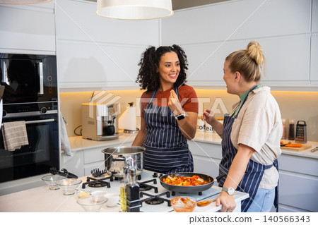 Diverse female friends in striped aprons cooking while chatting at kitchen island countertop Diverse female friends in striped aprons cooking while chatting at kitchen island countertop 140566343