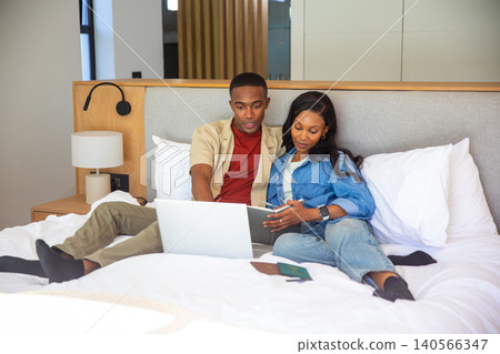 African American couple sitting on bed in bedroom planning trip on laptop with passports and tablet African American couple sitting on bed in bedroom planning trip on laptop with passports and tablet 140566347