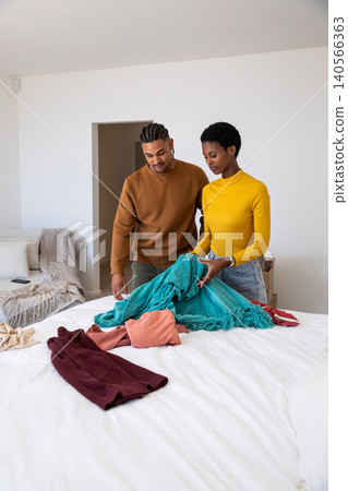 Couple folding teal blanket, burgundy sweater and peach shirt on white duvet in bedroom with sofa Couple folding teal blanket, burgundy sweater and peach shirt on white duvet in bedroom with sofa 140566363