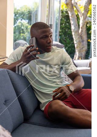 African american man holding smartphone, wearing watch, sitting on sofa by glass door African american man holding smartphone, wearing watch, sitting on sofa by glass door 140566562