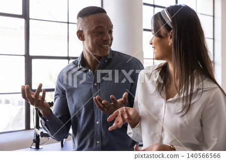 Diverse coworkers standing by grid windows, white column, gesturing over desk lamp, monitor Diverse coworkers standing by grid windows, white column, gesturing over desk lamp, monitor 140566566