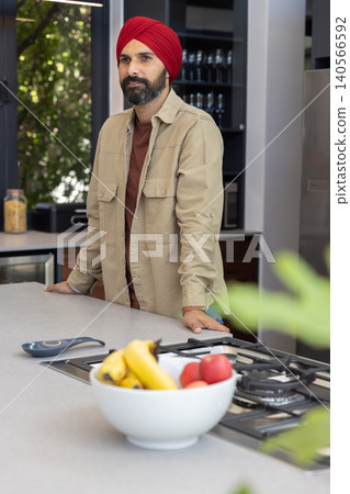Mid adult indian man in red turban standing behind kitchen island by fruit bowl, stovetop Mid adult indian man in red turban standing behind kitchen island by fruit bowl, stovetop 140566592