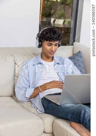 Asian male wearing headphones sitting on sofa using laptop in living room near patio door Asian male wearing headphones sitting on sofa using laptop in living room near patio door 140566597
