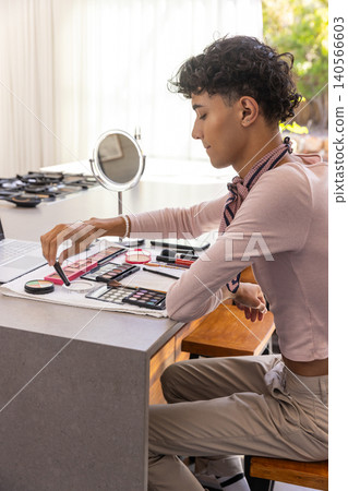 Man applying eyeshadow on makeup palettes at kitchen table with laptop and mirror, copy space 140566603