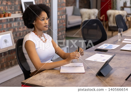 African american woman wearing top taking notes in notebook at conference table with tablet African american woman wearing top taking notes in notebook at conference table with tablet 140566654