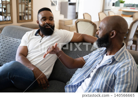 African American male friends chatting casually on gray sofa in living room with dining table African American male friends chatting casually on gray sofa in living room with dining table 140566665