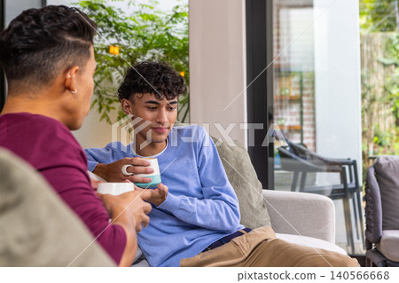 Male friends sipping coffee from ceramic mugs on sofa with throw pillow next to sliding glass door Male friends sipping coffee from ceramic mugs on sofa with throw pillow next to sliding glass door 140566668