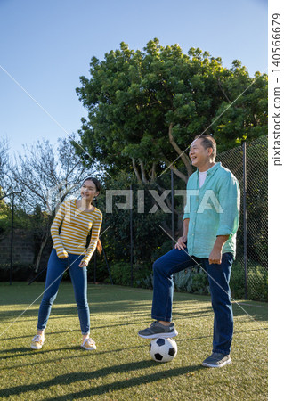 Asian couple standing in grassy yard with foot resting on soccer ball next to chain-link fence Asian couple standing in grassy yard with foot resting on soccer ball next to chain-link fence 140566679
