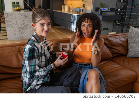 Female couple sitting on sofa next to marble countertop holding red jewelry box expressing surprise Female couple sitting on sofa next to marble countertop holding red jewelry box expressing surprise 140566702