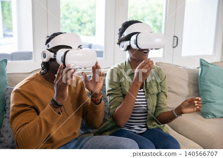 African american couple wearing vr headsets raising and gesturing on couch in living room African american couple wearing vr headsets raising and gesturing on couch in living room 140566707