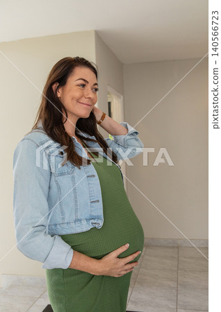 Mid adult pregnant woman standing in house hallway cradling belly while adjusting denim jacket Mid adult pregnant woman standing in house hallway cradling belly while adjusting denim jacket 140566723