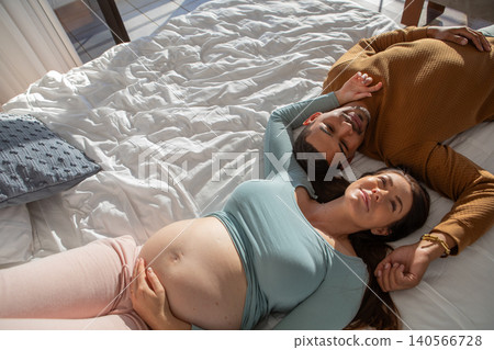 Diverse female couple lying on white comforter in sunlit bedroom holding hands and touching belly Diverse female couple lying on white comforter in sunlit bedroom holding hands and touching belly 140566728