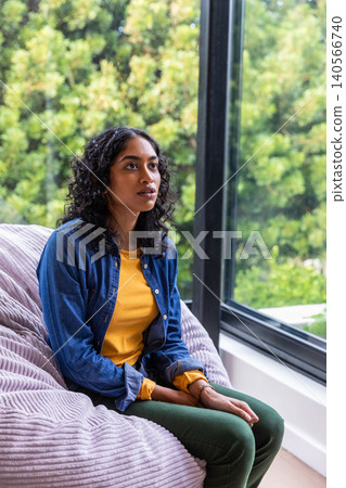 Asian woman sitting on beanbag at home by window overlooking foliage, copy space Asian woman sitting on beanbag at home by window overlooking foliage, copy space 140566740