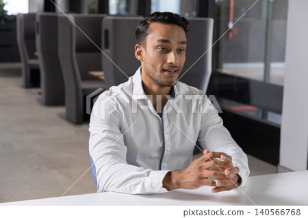 Asian man in shirt sitting and listening at white table in office lounge with glass partition Asian man in shirt sitting and listening at white table in office lounge with glass partition 140566768