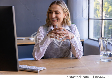Woman smiling holding glass of water and typing on wireless keyboard at wooden desk Woman smiling holding glass of water and typing on wireless keyboard at wooden desk 140566792