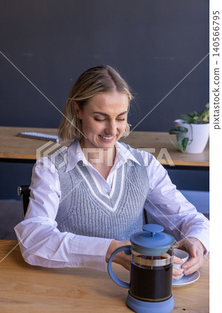 Woman pouring coffee from french press into cup at office desk with keyboard Woman pouring coffee from french press into cup at office desk with keyboard 140566795