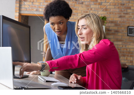 Diverse female coworkers collaborating at office desk with laptop and external monitor Diverse female coworkers collaborating at office desk with laptop and external monitor 140566810
