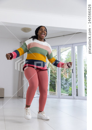 African american female jumping in living room next to patio doors wearing sweater and sneakers African american female jumping in living room next to patio doors wearing sweater and sneakers 140566826