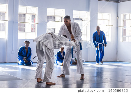 Male martial arts students grappling in bright dojo on padded mats with gis and belts Male martial arts students grappling in bright dojo on padded mats with gis and belts 140566833