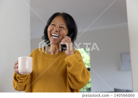 Senior Asian woman smiling while holding smartphone and white mug at home wearing mustard sweater Senior Asian woman smiling while holding smartphone and white mug at home wearing mustard sweater 140566860