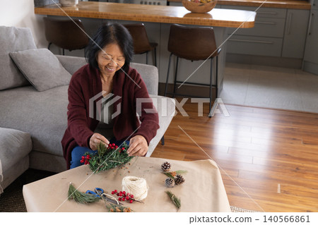 Senior Asian woman creating holiday wreath on gray sofa at home with pine cones and berries Senior Asian woman creating holiday wreath on gray sofa at home with pine cones and berries 140566861