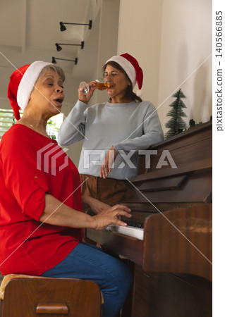 Diverse senior female friends singing, sipping from flute at piano in music room wearing Santa hats Diverse senior female friends singing, sipping from flute at piano in music room wearing Santa hats 140566885