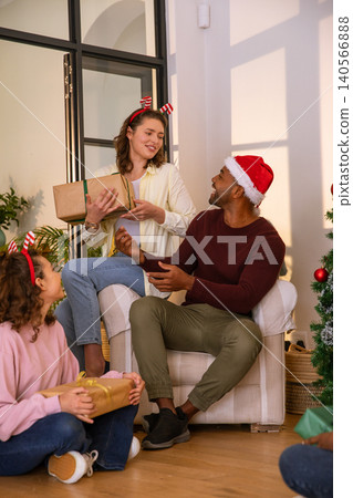 Diverse family exchanging wrapped gifts, wearing santa hat, headbands by christmas tree at home Diverse family exchanging wrapped gifts, wearing santa hat, headbands by christmas tree at home 140566888