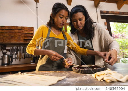 Asian mother and Hispanic daughter weaving pastry strips over cherry pie on countertop in aprons Asian mother and Hispanic daughter weaving pastry strips over cherry pie on countertop in aprons 140566904