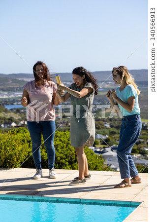 Diverse female friends standing, popping party popper on pool deck beside rectangular pool 140566913
