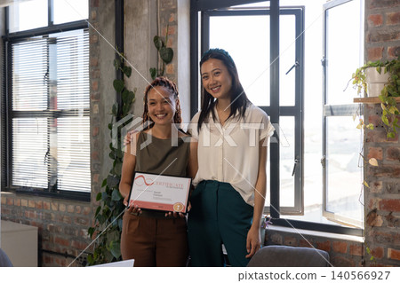 Diverse female colleagues in business attire standing in office holding framed certificate Diverse female colleagues in business attire standing in office holding framed certificate 140566927