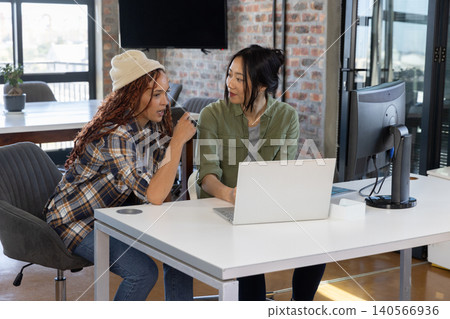 Diverse female coworkers collaborating on laptop and monitor at white desk in open-plan office Diverse female coworkers collaborating on laptop and monitor at white desk in open-plan office 140566936