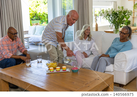Diverse senior friends pouring water from pitcher sitting around coffee table in living room Diverse senior friends pouring water from pitcher sitting around coffee table in living room 140566975