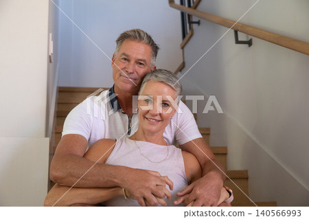 Senior couple sitting on wooden staircase in hallway embracing by handrail and light walls Senior couple sitting on wooden staircase in hallway embracing by handrail and light walls 140566993