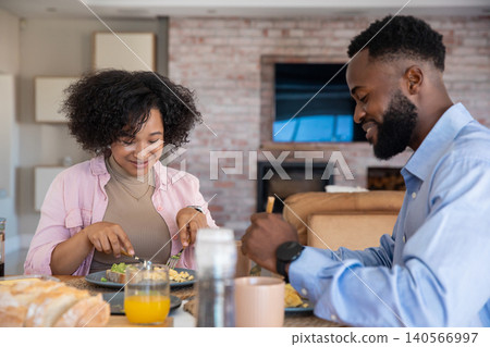African american couple sitting at dining table cutting breakfast food, sipping orange juice African american couple sitting at dining table cutting breakfast food, sipping orange juice 140566997
