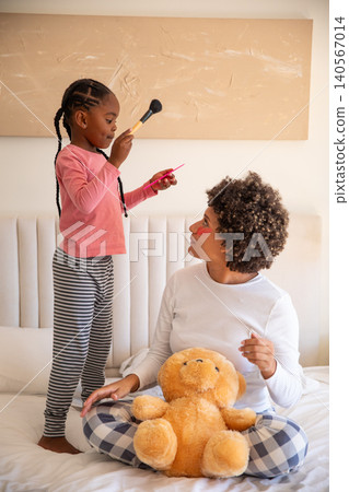 Diverse mother with daughter playing makeup on bed in bedroom with makeup brush and lip gloss Diverse mother with daughter playing makeup on bed in bedroom with makeup brush and lip gloss 140567014