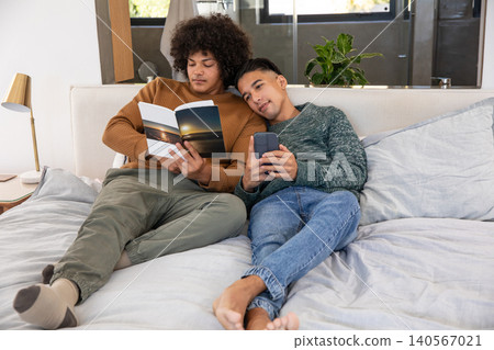 Two men reclining on gray duvet reading sunset paperback book while checking smartphone in bedroom Two men reclining on gray duvet reading sunset paperback book while checking smartphone in bedroom 140567021