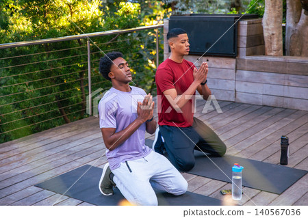 Diverse male friends meditating while kneeling on yoga mats on deck near water bottles smartphone 140567036