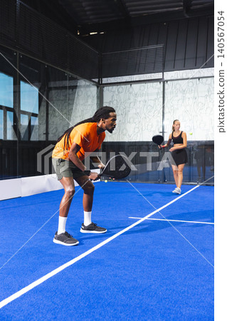 Diverse sports partners holding rackets and tracking ball on blue padel court, copy space Diverse sports partners holding rackets and tracking ball on blue padel court, copy space 140567054